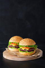Two homemade burger on little wooden cutting board over dark background.