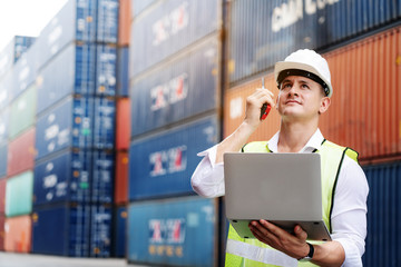 Engineer or foreman use walkie-talkie and laptop. In order to move container and shipping. Safety helmet help prevent accident from various equipment in factory. Industry and Transportation concept - obrazy, fototapety, plakaty