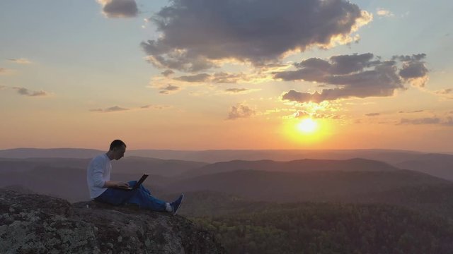 Epic drone shot of a silhouette of a young freelancer with a laptop on the edge of a rock ledge in the mountains.