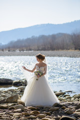 The bride plays with a veil on a mountain river. Holding a wedding bouquet in his hands.