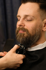 Barber trimming beard of male customer with electric razor at barbershop. Man with beard and...