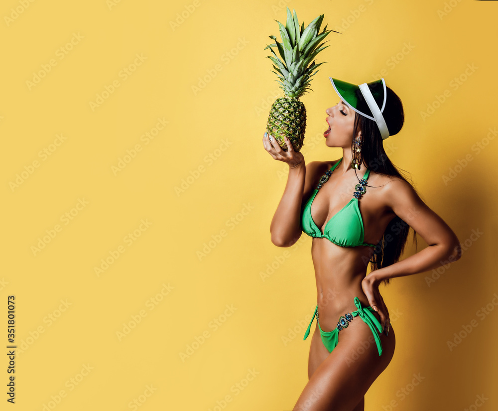 Wall mural young tanned slim brunette woman in sexy green bikini with decorations and visor is going to bite a 