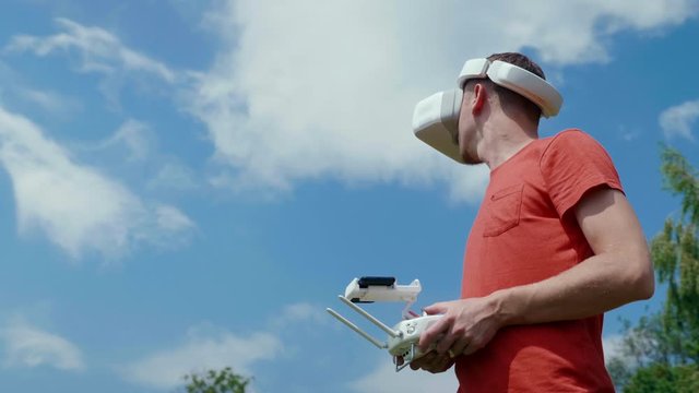 Man controls a quadrocopter through a remote control and looks at the video with goggles on his head. Drone operator in red T-shirt in a park shoots video on drone. Blue sky on the background