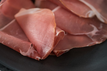 Thin italian prosciutto slices on black plate on conctere background closeup