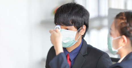 Male staff wearing medical face mask is checking fever by his colleague using infrared forehead thermometer inside the business office during coronavirus outbreak for new normal policy