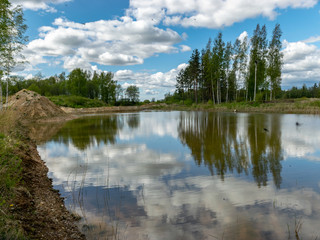 a fish pond, in the water of which there are colorful cumulus clouds and trees, there are recently dug gravel piles on the shore of the pond, the first spring greenery in nature