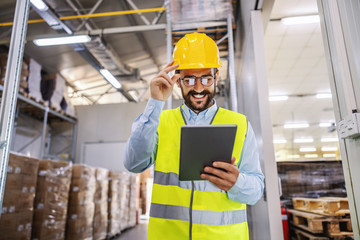Young smiling supervisor in vest and protective helmet standing in warehouse and using tablet for work.