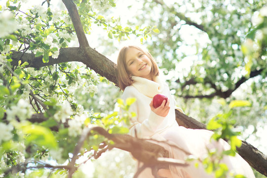 Happy beautiful teenage girl enjoying spring sunny day sitting on blossom tree ready to eat red delicious apple