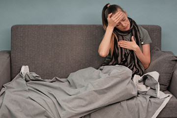 The girl is sick at home. Sitting on a couch, wrapped in a blanket, a scarf around his neck. Pain in the head, migraine. Coronavirus.