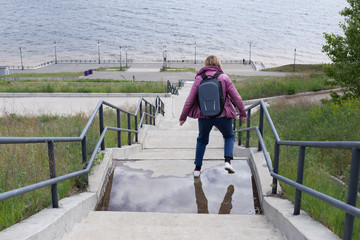 River embankment and high staircase. A woman is jumping through a puddle. A shot from the back high above the shore.
