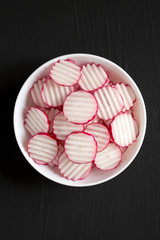 Fresh Radish Chips Slices in a white bowl on a black surface, top view. Flat lay, overhead, from above. Copy space.