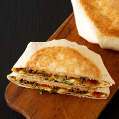 Tasty Homemade Beef Crunchwrap Quesadilla on a rustic board on a black background, close-up.