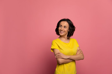 Pink and yellow. Close-up photo of attractive young girl in a yellow t-shirt, who is looking aside with folded arms and smiling broadly.