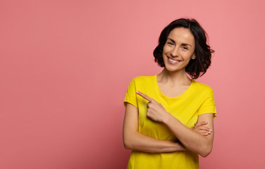 Fototapeta premium Pointing. Close-up photo of a happy woman in yellow t-shirt, who is looking in the camera and pointing to the left with her left index finger.