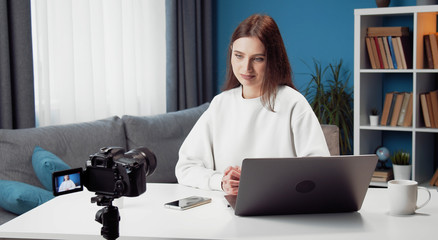 Young female vlogger recording video on camera sitting at desk in front of laptop at home