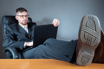 The man is sitting in a chair with his feet on the table. The man with the laptop put his feet up on the table. A businessman in a suit is sitting in a chair with a laptop.