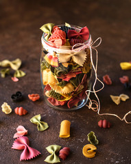colored italian pasta of different shapes in a glass jar on a brown texture background