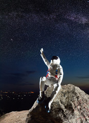 Space traveler sitting on top of rocky hill and raising hand while looking at beautiful starry sky....