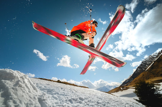 A Young Athlete Skier Does A Trick On A Snow Kicker Jump And Cross Skis In A Counter Light Against A Blue Sky And Clouds In The Mountains.