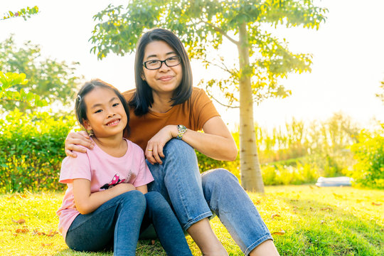 Asian mother and child daughter sitting on the grass slopes of the public gardens park. Mother holds daughter's shoulder. Both smile happily. Golden evening light covered the entire image.