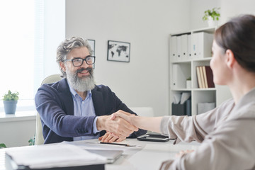 Smiling mature employer with grey beard and hair shaking hand of young female