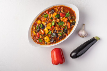 Homemade vegetable stew of eggplant, zucchini, tomatoes, peppers, garlic, prunes and greens on bowl on light background. Top view. Copy space for text