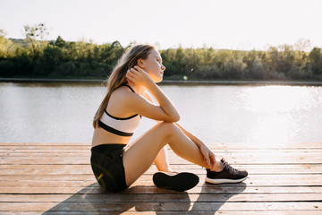 Young slim fit woman resting after workout, sitting on a wooden platform by the lake, outdoors. Sportive girl enjoying sun light and fresh morning air outside.