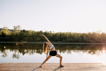 Fototapeta na wymiar Young slim fit woman doing yoga exercise, spine stretching pose, in the morning. Pilates instructor outdoors, on a wooden platform by the lake.