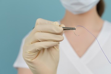 doctor holds a surgical thread with a needle in gloves with a mask on a blue background