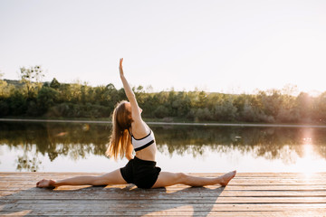 Young slim fit woman doing yoga exercise, twine pose, in the morning. Pilates instructor outdoors, on a wooden platform by the lake.