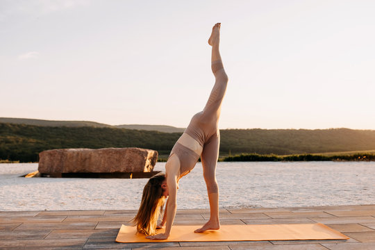 Young slim fit woman on a sport mat standing in yoga split pose at the sunset, outdoors. Sportive girl doing pilates.