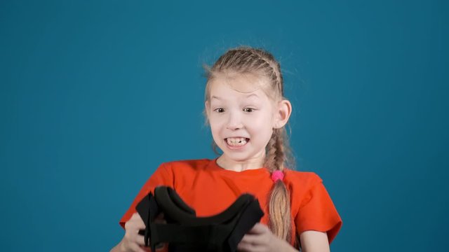 cute little child with long braid takes off virtual reality headset scared by screen picture on blue background slow motion