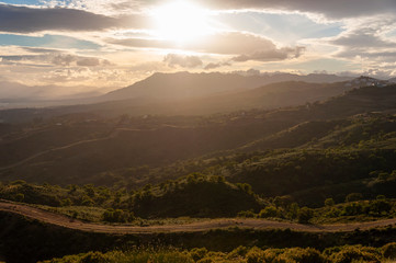 Bright sunshine during sunset over the mountain peaks and a country road with green hills in the foreground.