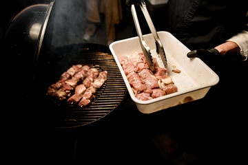 person with tongs spread raw meat pieces on barbecue grill