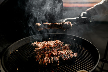 male chef's hand using tongs holds piece of meat over barbecue.