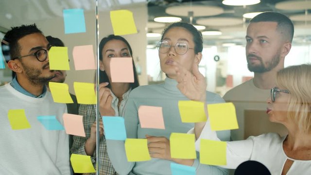 Young creative men and women are working with sticky notes on glass board discussing ideas solving problems concentrated on mind map in office.