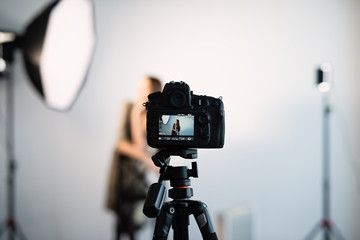 the camera stands on a tripod, a picture is displayed on the screen, the model poses in front of the camera, a girl with brown hair.