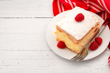 Authentic European dessert concept with piece of vanilla and soft custard cream cake (German: Cremeschnitte, Polish: Kremowka)and raspberries on white wood background and red tea cloth with copy space