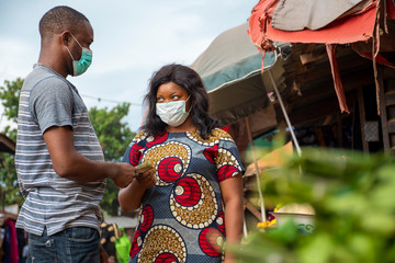 an african woman in a local market wearing a face mask selling to a customer taking money