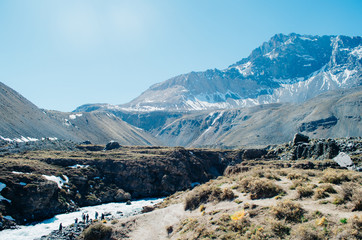 Rocky trekking path of the Andes Mountains with a group of people near a river and with a huge mountain partially covered in snow in the background with clear blue skies and a sun flare.