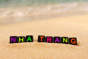The word Nha Trang is made up of colored letters on black cubes on sand with a blurry sea in the background