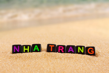 The word Nha Trang is made up of colored letters on black cubes on sand with a blurry sea in the...