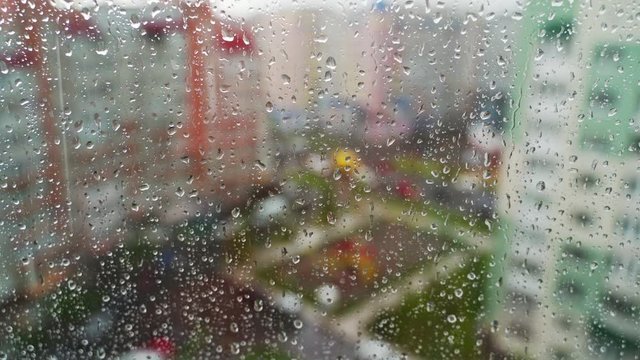Wet window on blurred city background. Raindrops moving down on glass window at rainy weather. Water droplets on window pane.