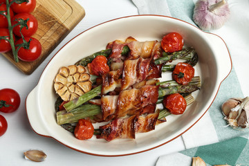Oven baked asparagus wrapped with bacon in ceramic dish on white wooden table, flat lay