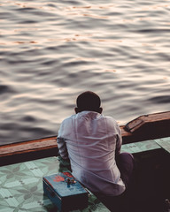 An unidentified boat operator gazing into the distance at Dubai Creek during a beautiful blue hour. Man sitting in an anchored wooden boat with a money box beside him.