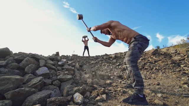 Sportsmen doing physical exercises on rocky hill. Strong man splits stones with a big hammer on a background of athlete who is throwing stones. Slow motion.