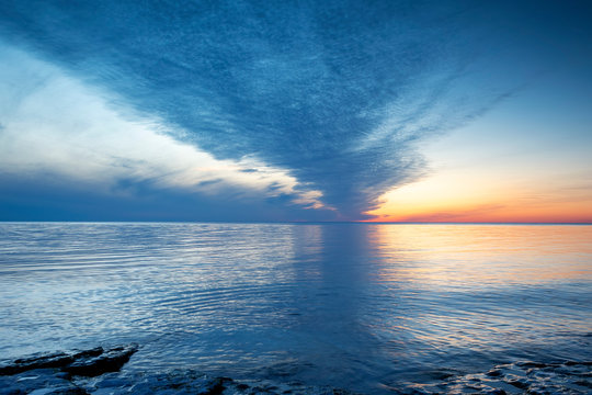 Cloudy summer sunset reflecting in ocean with endless horizon and deep blue ocean, silhouette of boulders laying in the foreground in shallow water at island of Gotland, Sweden