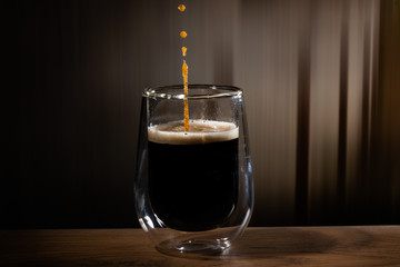 Coffee splash. Reaction of a falling drop on the hot, steaming and frothy coffee surface in a modern glass transparent cup. Studio photo with matching colored background