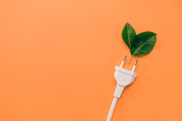 Creative top view flat lay of electrical plug, fresh green leaves with copy space on orange background in minimal style. Save energy concept.