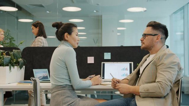 Asian woman and Caucasian man coworkers are talking in open space office sitting at desk discussing work with serious faces. People and workplace concept.